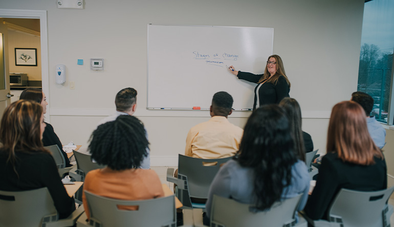 people in class listening to a teacher pointing at a white board
